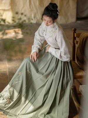 Side view of a woman wearing a sage green pleated Mamianqun Hanfu skirt and embroidered long-sleeve top while seated in a cozy traditional Chinese-inspired room