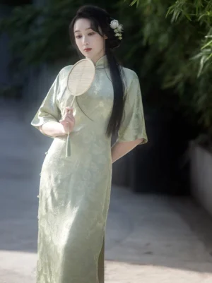 Sage green modern Chinese style cheongsam dress with floral jacquard fabric, classic side frog buttons, retro flared sleeves, and round bamboo fan
