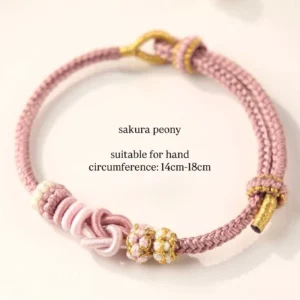 Sakura peony handwoven Chinese knot bracelet in dusty pink braided cord with gold tone accents, pale pink beads, and traditional knot decorations on a white background