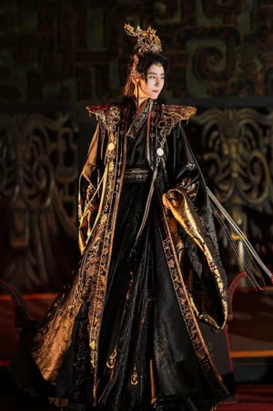 Front three quarter view of a male model wearing the black and gold Sanxingdui Star Priest xianxia hanfu costume with wide sleeves, ornate golden shoulder armor, embroidered robes, and long pearl tassels