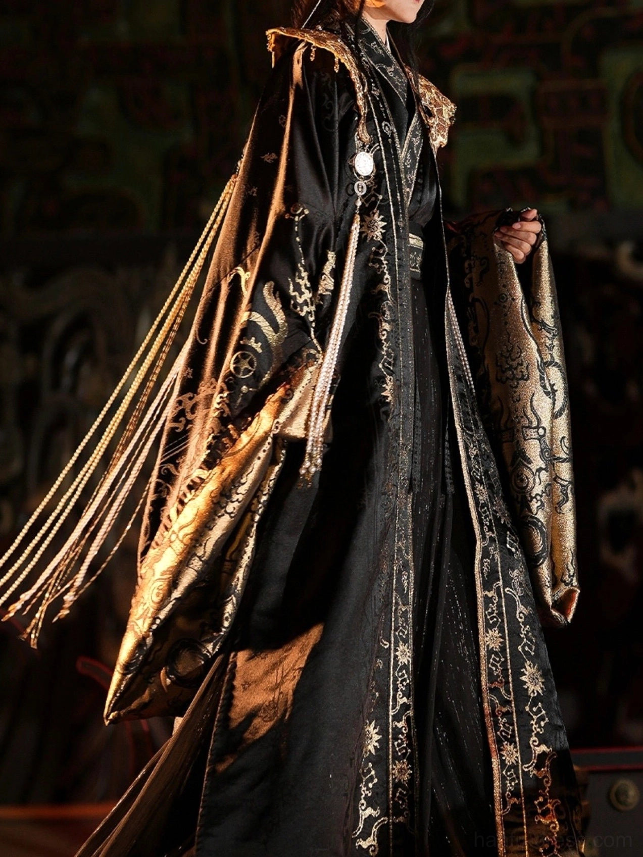 Back three quarter view of the Star Priest male hanfu costume showing shimmering black satin fabric, gold embroidered robe hem, wide sleeves, shoulder ornament, and cascading ceremonial tassels