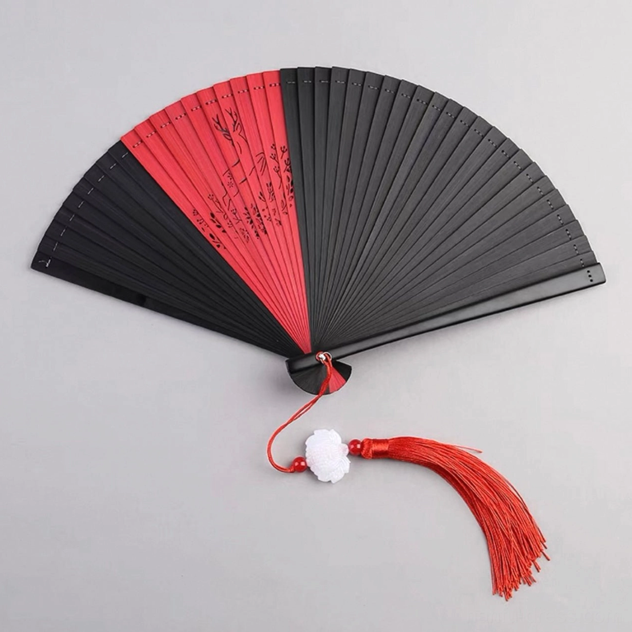 Scarlet red and black Chinese folding hand fan with calligraphy-inspired patterns, bamboo ribs, white flower charm, and red tassel on a light gray background