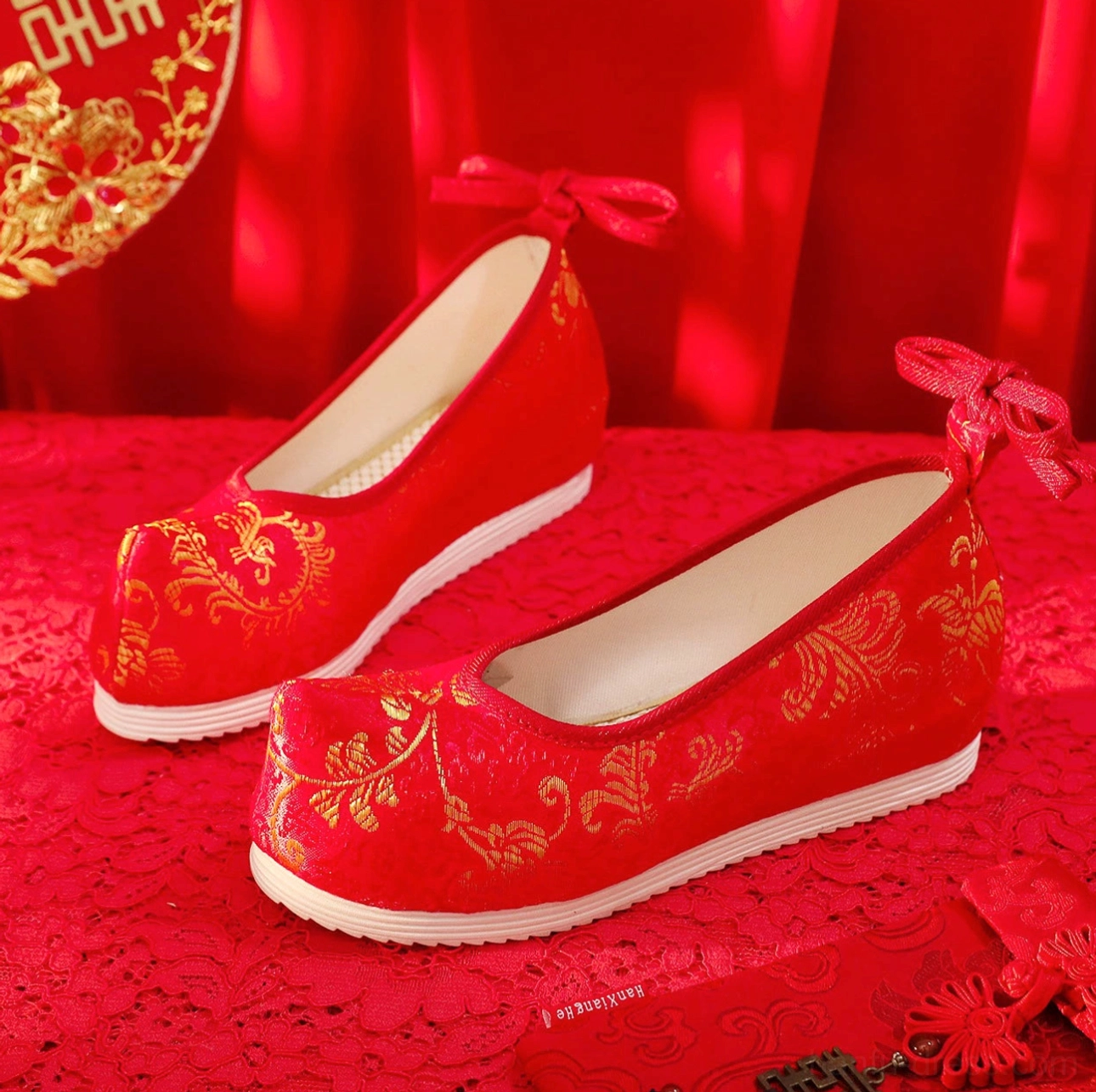 Scarlet traditional Chinese bridal Hanfu shoes with golden floral embroidery, upturned toe, red heel bow, and white rubber sole displayed on a Chinese wedding backdrop