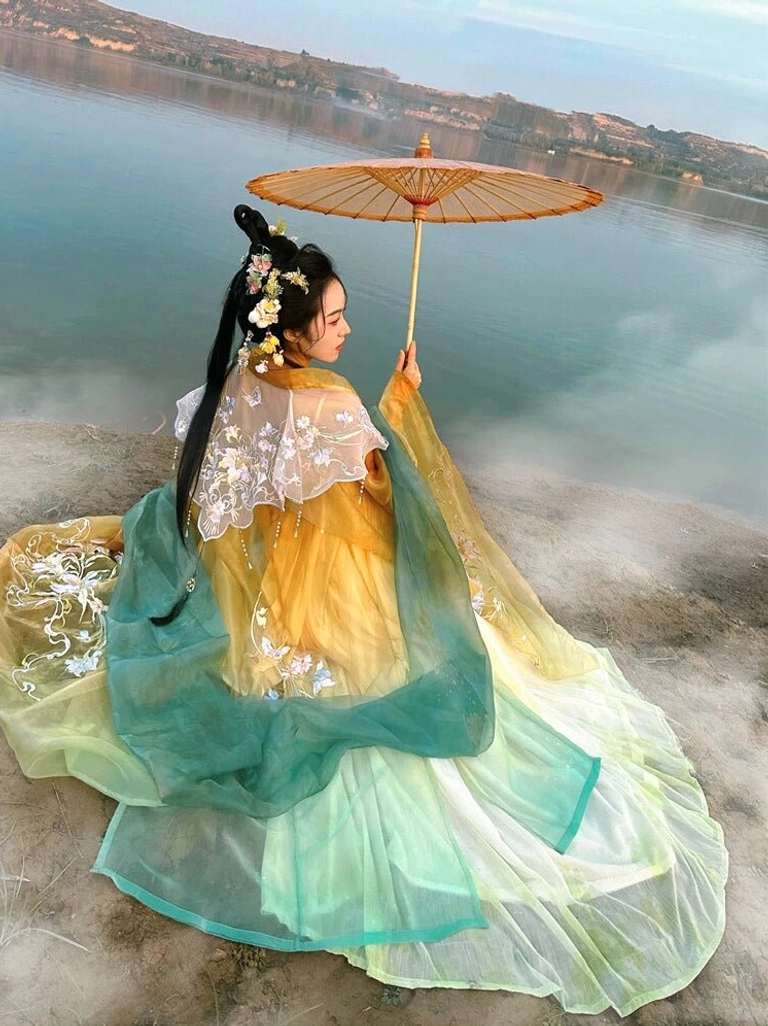 Seated outdoor model wearing a flowing Tang Dynasty Hanfu Heziqun dress with floral embroidery gradient fabric cyan accents and traditional paper umbrella
