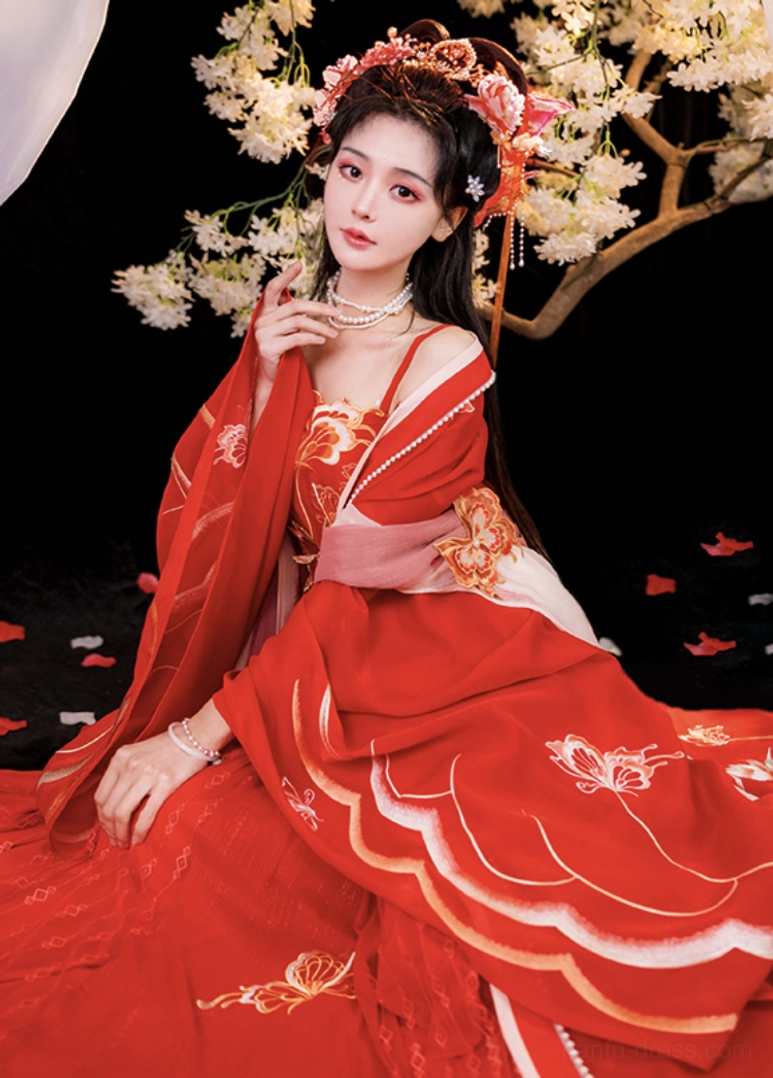 Seated view of a red traditional Chinese bridal hanfu showing scalloped embroidered hem, golden butterfly motifs, decorated camisole bodice, wide sleeves, and pearl necklace styling