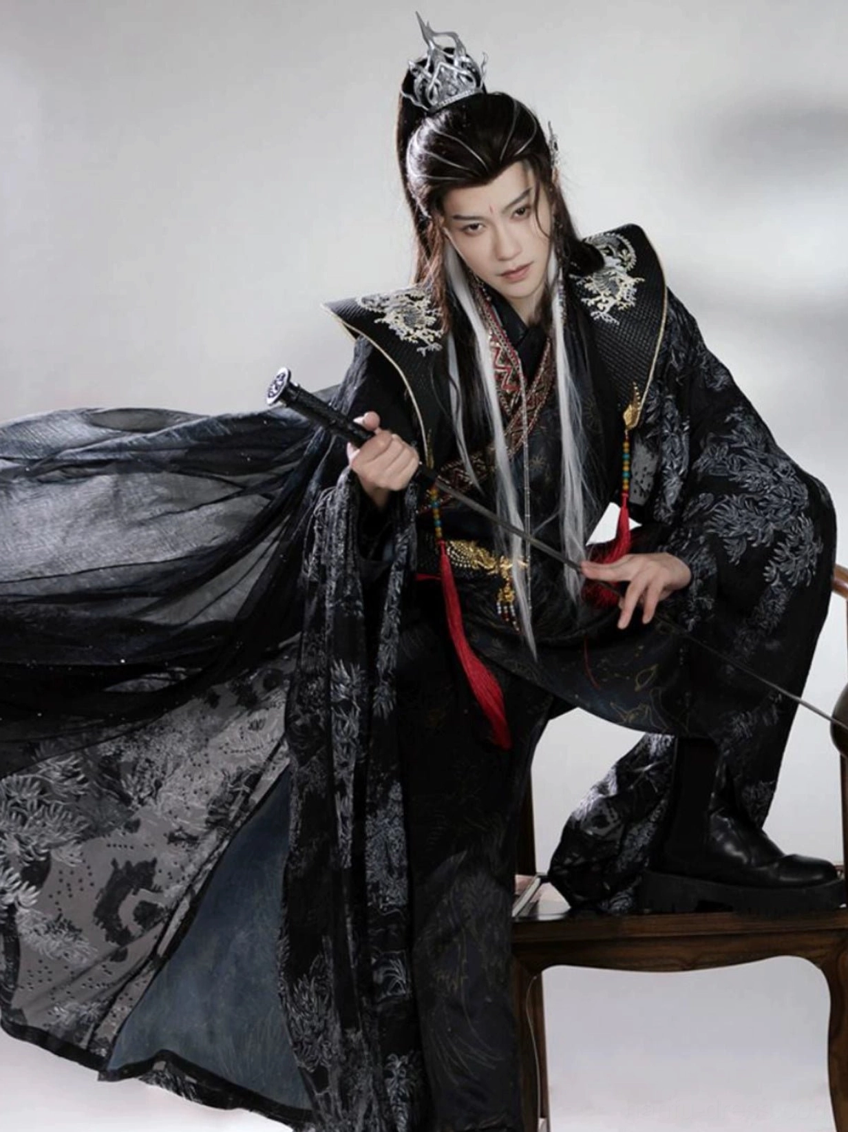 Dark Xianxia Hanfu for men styled in a dramatic seated swordsman pose with layered black robe, sheer cape, wide sleeves, and red tassel belt