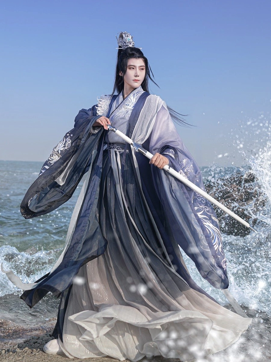 Front full-body view of Shadowborne Blade Cheng Ying Jian wuxia hanfu for men with blue gradient layered robes, phoenix embroidery, flowing sleeves, and ceremonial sword beside the sea