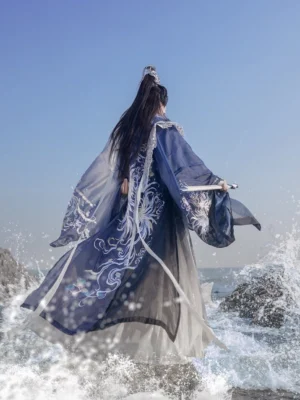Back view of Shadowborne Blade male hanfu standing on seaside rocks, showing the flowing blue gradient hem, embroidered wide sleeves, and layered wuxia robe silhouette