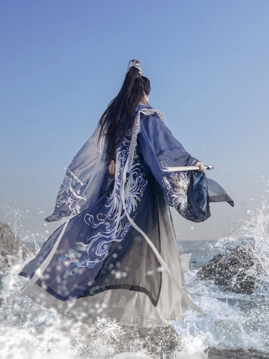 Back view of Shadowborne Blade male hanfu standing on seaside rocks, showing the flowing blue gradient hem, embroidered wide sleeves, and layered wuxia robe silhouette
