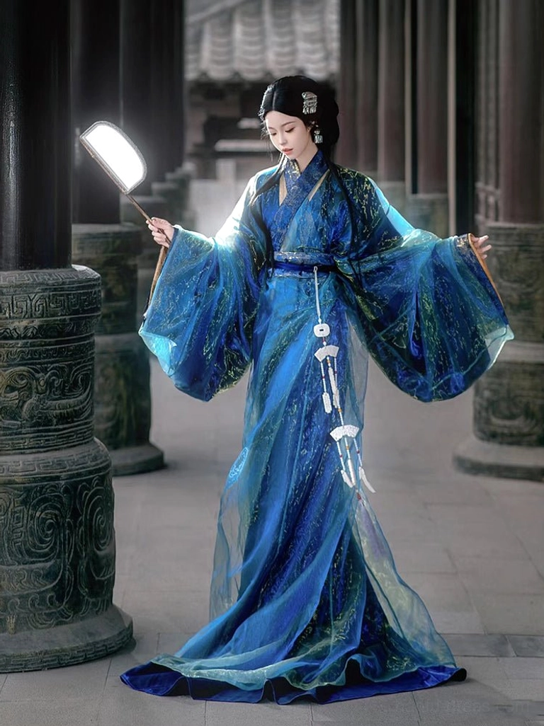Full-length three-quarter view of a woman wearing a shimmering deep blue Warring States and Han Dynasty straight-hem Hanfu robe with wide flowing sleeves and white jade-style tassel ornaments in an ancient Chinese courtyard