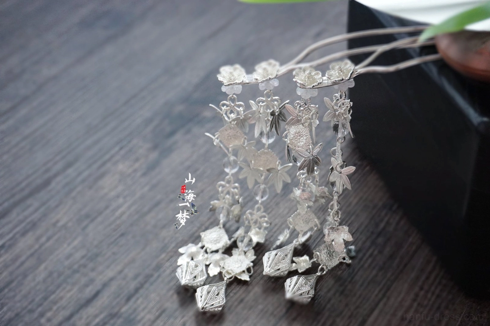 Pair of silver traditional Chinese hairpin designs with intricate filigree flowers, geometric pendants, and dual hairpin sticks on a wooden display