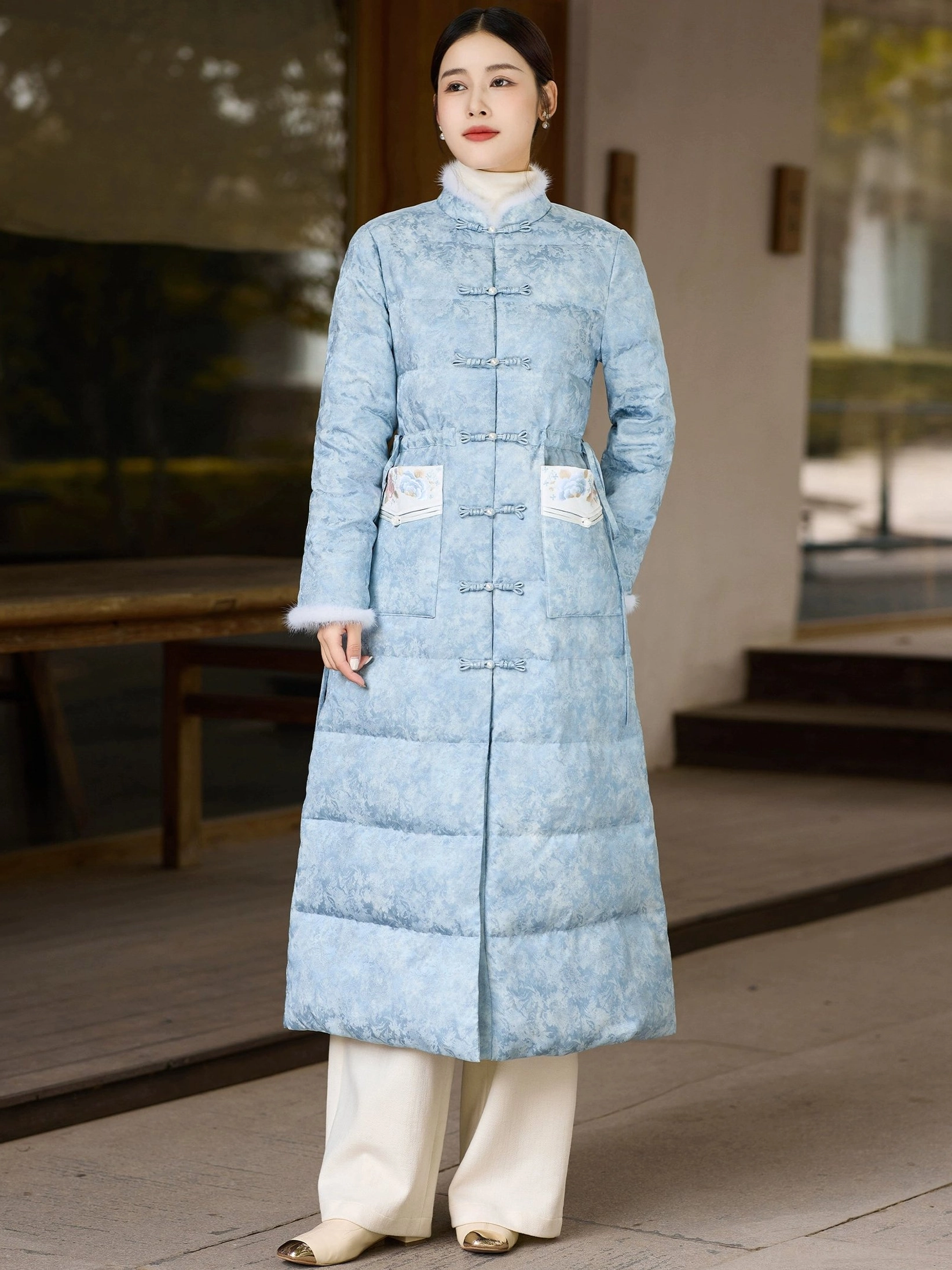 Back three-quarter view of women's long sky blue Hanfu-inspired winter down coat with quilted panels plush cuffs and embroidered pocket accents