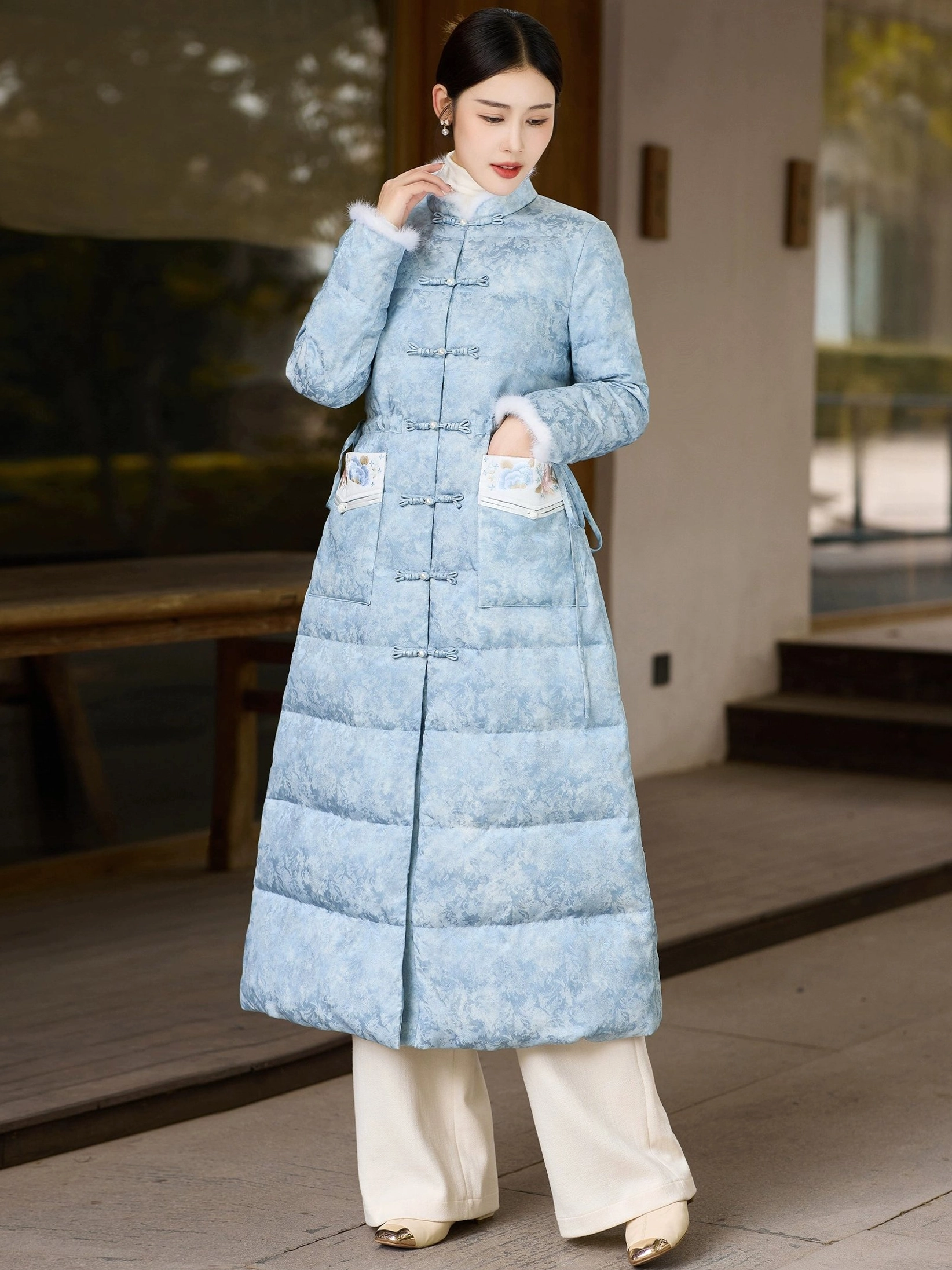Right three-quarter view of sky blue qipao-inspired women's down overcoat with stand collar full frog button closure and embroidered patch pockets