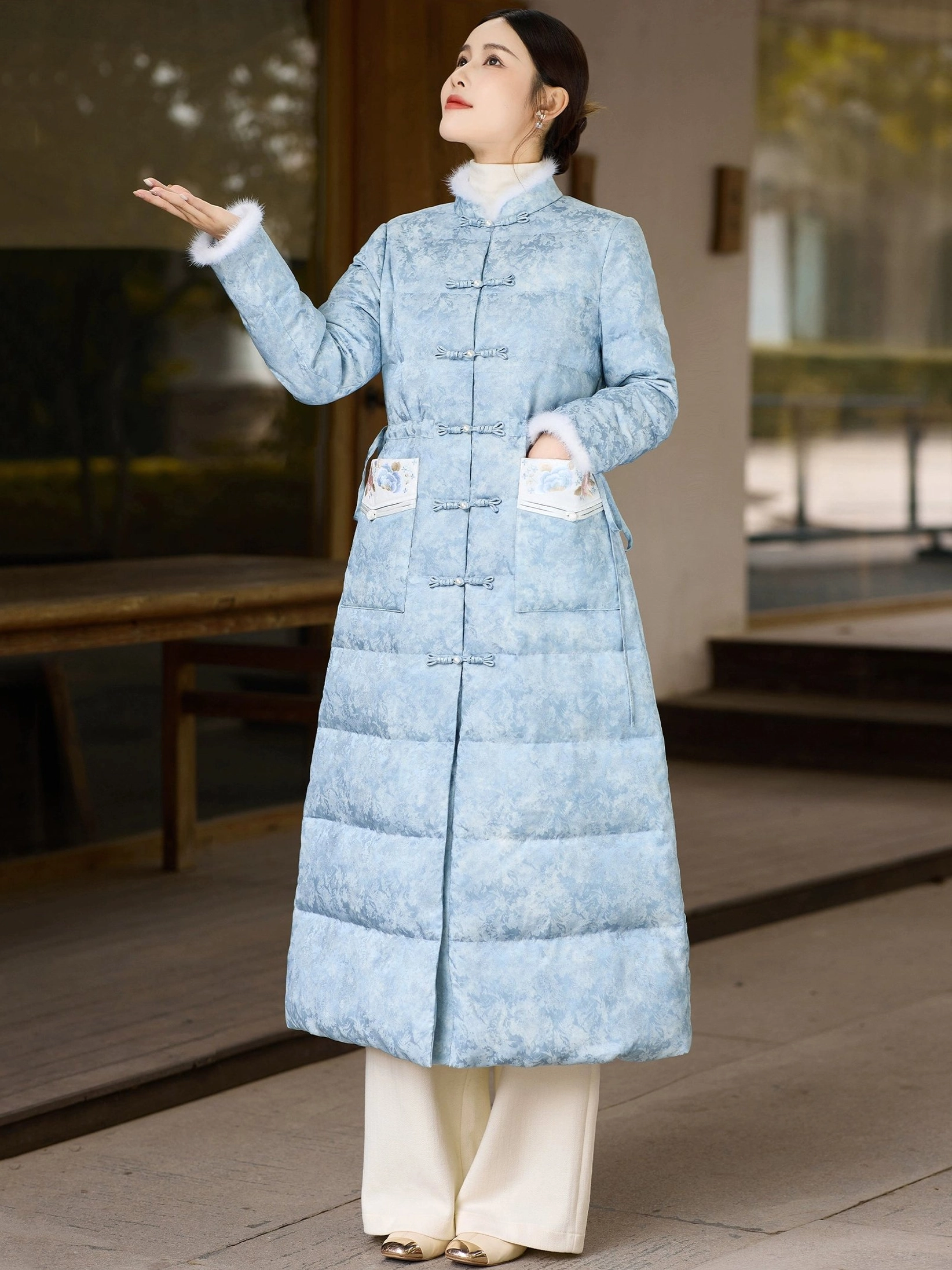 Three-quarter side view of women's sky blue Chinese style long down coat showing plush cuff trim quilted insulation and embroidered pocket details
