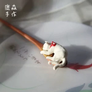 Top-down close-up of a handmade sleeping cat Chinese hair stick with a warm brown wooden shaft, white cat ornament, red bow, and golden cushion detail for Hanfu hairstyles