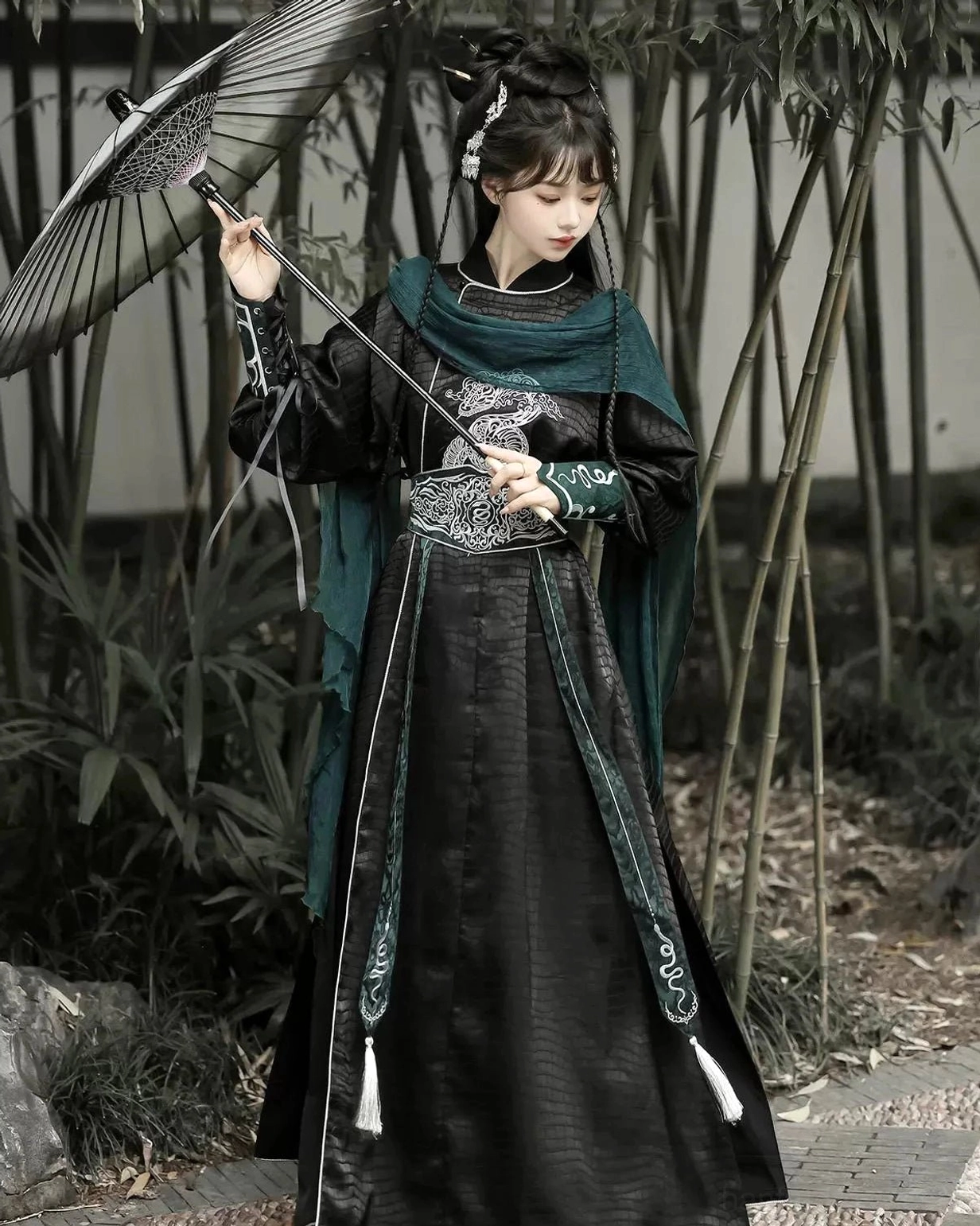 Female warrior wearing a black Snake Encounter xianxia Hanfu costume with silver snake embroidery, dark green scarf, and traditional Chinese paper umbrella in a bamboo courtyard