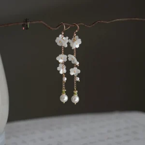 Snow bloom Chinese earrings with white floral charms, pearl drops, green beads, and gold toned details displayed on a natural twig