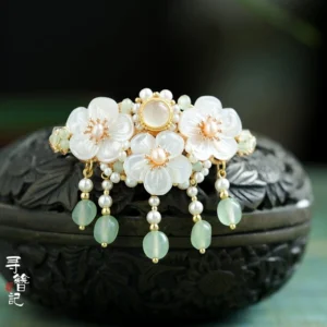 Snow Petals Chinese Hanfu back hair pin displayed on a carved dark wooden jewelry box with white flower petals, cat-eye-style center gem, gold-toned frame, pearl tassel, and pale green drop bead
