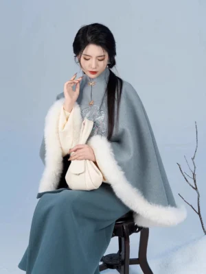 Seated model wearing a Song Dynasty hanfu outfit with embroidered dusty blue cape, teal wrap skirt, beige inner garment, and soft drawstring handbag