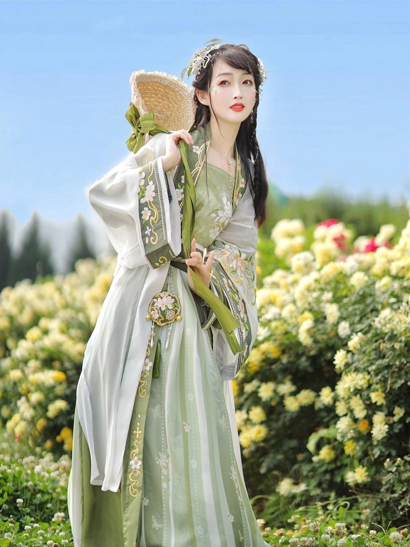 Side full-body view of a woman wearing green and ivory Song Dynasty hanfu with embroidered white beizi coat, waist pendant, and flowing skirt in a sunlit flower field