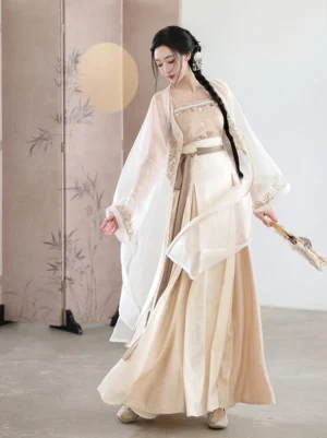 Three-quarter view of a cream and beige Song Dynasty-inspired Hanfu three-piece set with pleated wrap skirt, sheer embroidered cardigan, camisole, folding fan, and Chinese studio backdrop