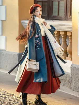 Front three-quarter view of a Song Dynasty-inspired Hanfu outfit featuring a dark teal embroidered beizi wool-look coat, burgundy pleated wrap skirt, off-white cross-collar top, red beret, scarf, and leather handbag on a European street