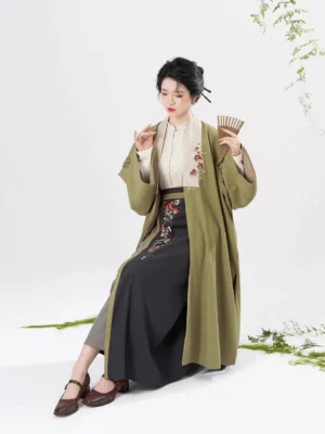 Seated model wearing complete Song Dynasty hanfu set with olive green outer coat embroidered skirt and folding fan