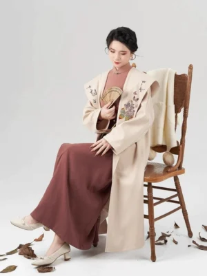 Seated woman wearing Song Dynasty inspired modern Hanfu outfit with hooded cream coat embroidered wrap skirt and folding fan