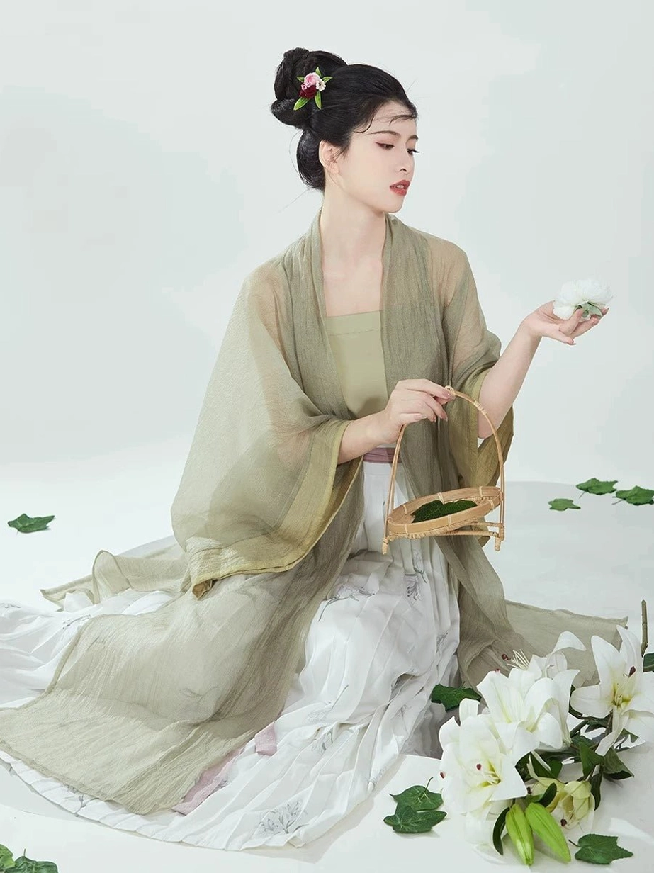 Side view of a woman kneeling in a flowing Song Dynasty Hanfu outfit with sage green robe, printed pleated skirt, bamboo tea basket, and floral styling