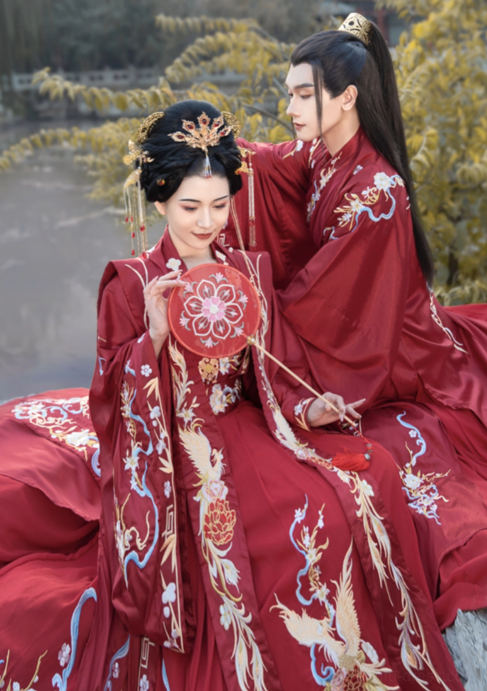 Crimson Chinese wedding hanfu couple by riverside showing phoenix embroidery and embroidered round bridal fan