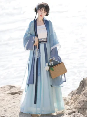 Front view of a Song Dynasty style Qiyao Ruqun Hanfu with blue to light cyan gradient pleated skirt, dark blue trimmed beizi robe, embroidered waist belt, and woven floral handbag beside a lakeside