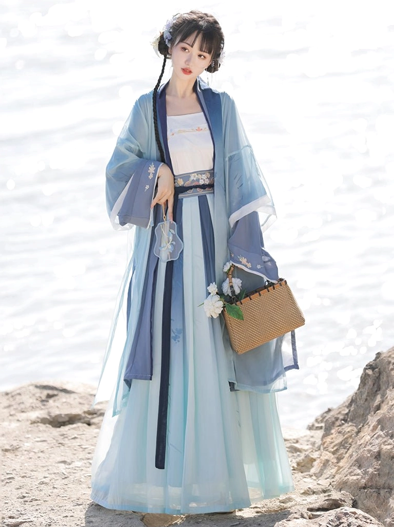 Front view of a Song Dynasty style Qiyao Ruqun Hanfu with blue to light cyan gradient pleated skirt, dark blue trimmed beizi robe, embroidered waist belt, and woven floral handbag beside a lakeside