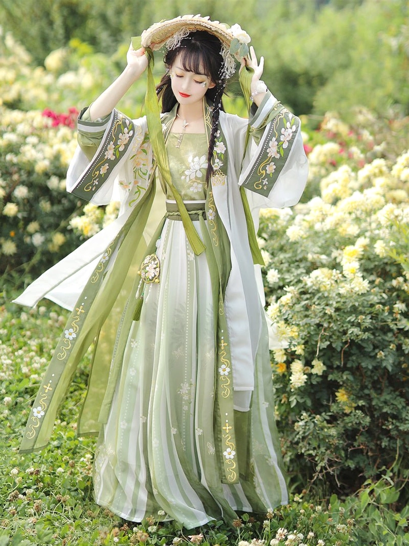 Front full-body view of a woman wearing a Song Dynasty qiyao ruqun hanfu set in soft green and ivory with floral embroidery, striped skirt, and wide-sleeved beizi coat in a flower garden