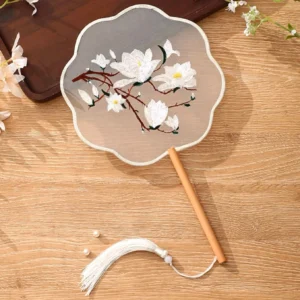 Traditional Chinese tuanshan round hand fan with scalloped edge, translucent embroidered silk face, white magnolia flower and branch design, wooden handle, and matching tassel displayed on a light wood table