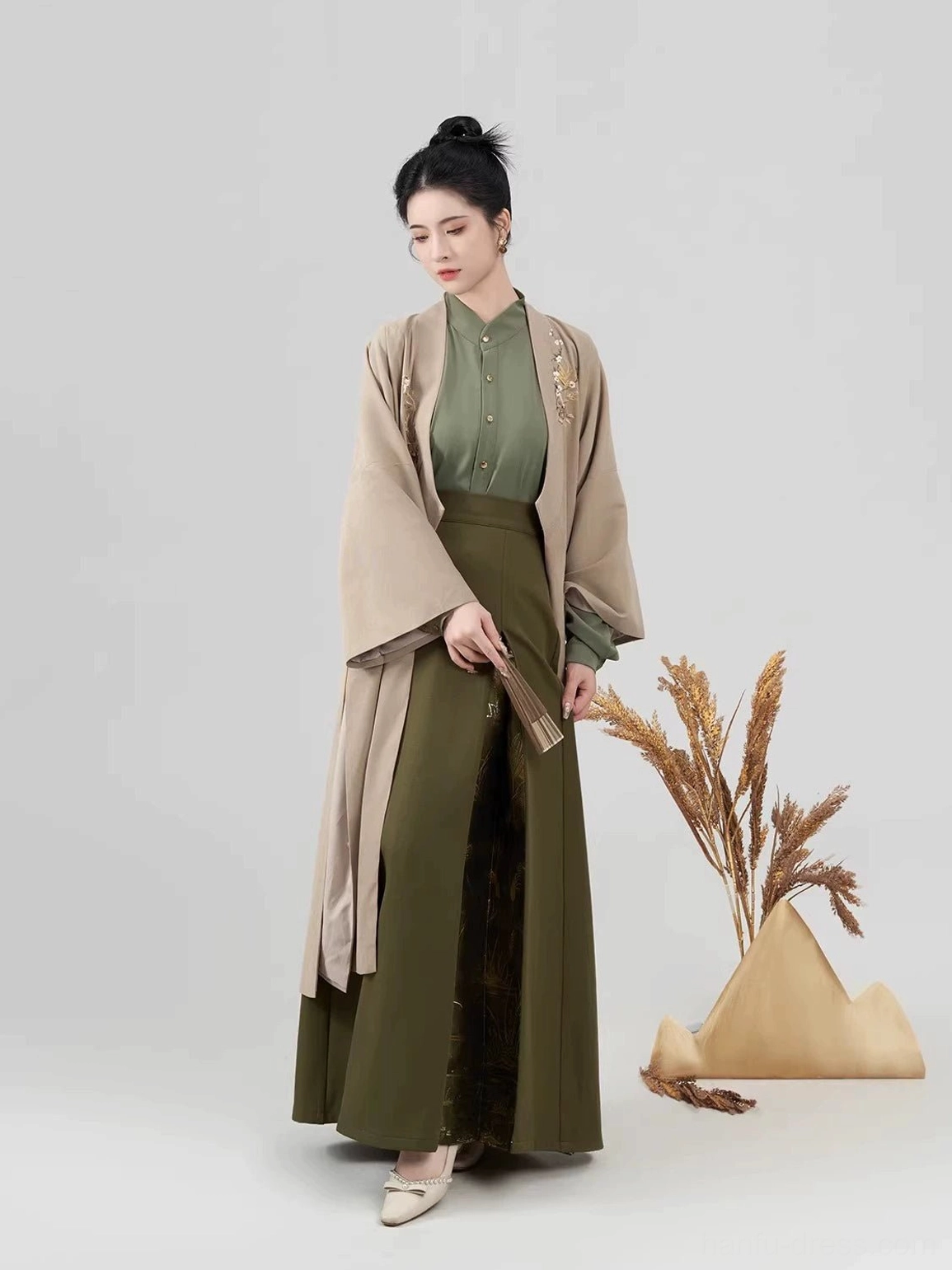 Side view of a woman wearing a flowing light khaki Song-style Hanfu coat over an army green top and pleated skirt while holding a traditional folding fan