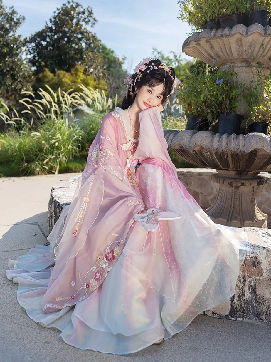 Seated model wearing Spring Banquet Tang Dynasty Hanfu heziqun with floral embroidery, soft pink gradient skirt, blue trailing sash, and flower hair ornaments