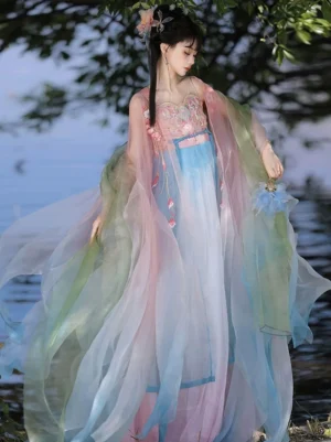 Front three-quarter view of a woman wearing a Spring Flowers Tang Dynasty Hanfu Heziqun dress with pastel pink blue white gradient skirt, floral embroidered bodice, and flowing sheer sleeves beside a lakeside