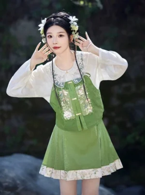 Model wearing a modern Ming-style Hanfu three-piece outfit with ivory lace-collar blouse, sage-green embroidered bustier, pleated floral mini skirt, and traditional Chinese hair ornaments beside a waterside landscape
