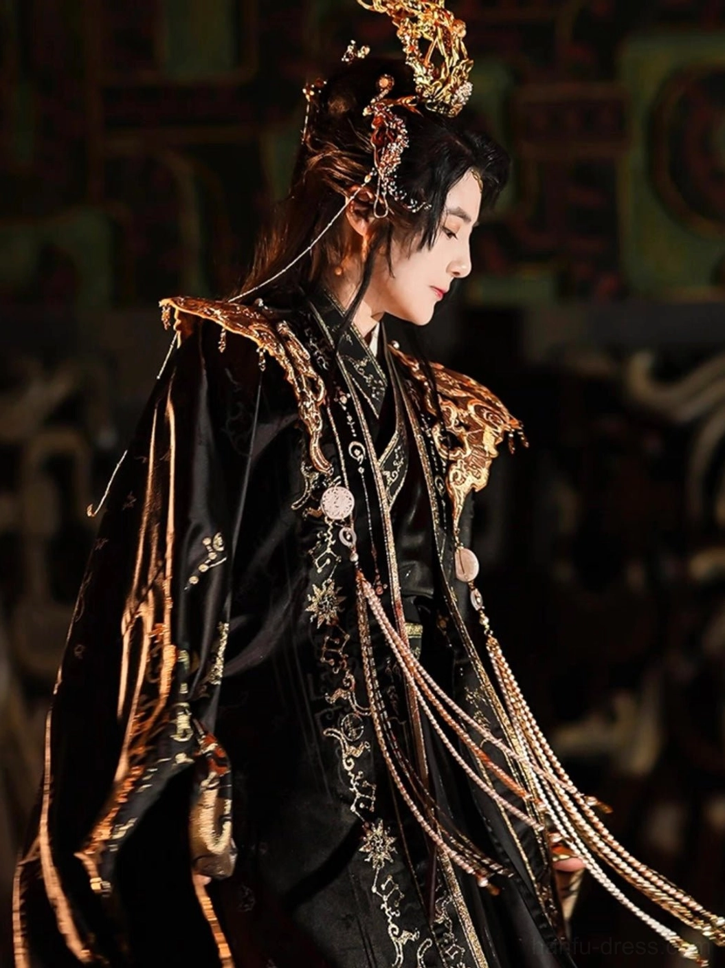 Side view of a male model in the black Star Priest hanfu costume featuring gold floral embroidery, layered beaded shoulder tassels, golden sleeve cuffs, and an elaborate ceremonial crown