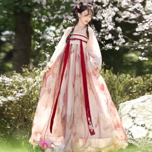 Front view of a pastel pink strawberry print Tang-style Hanfu dress in a cherry blossom woodland, showing elegant lantern sleeves, a long flowing skirt, and contrasting red decorative ribbons