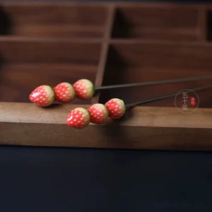 Two strawberry themed Chinese hair sticks for Hanfu shown on a dark tabletop with wooden storage tray, each decorated with three detailed strawberry beads