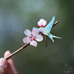 Close-up of a handmade Chinese hair stick with a blue swallow, golden decorative lines, pink peach blossoms, and a brown wooden shaft held against blurred green foliage