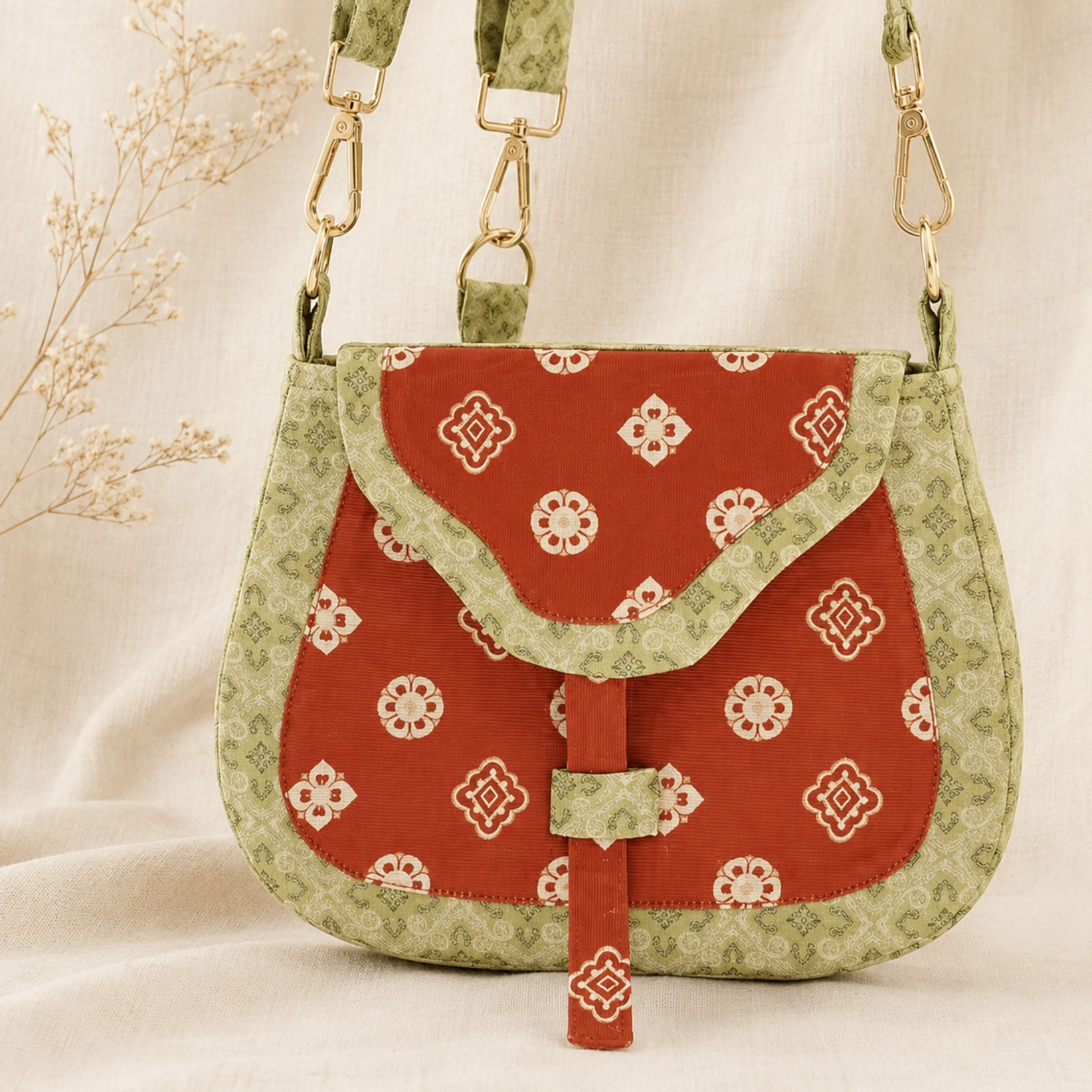 Tang Dynasty floral Chinese canvas crossbody bag shown from the front on linen fabric, featuring a red saddle-shaped body, beige traditional floral motifs, green patterned brocade trim, and gold-tone hardware