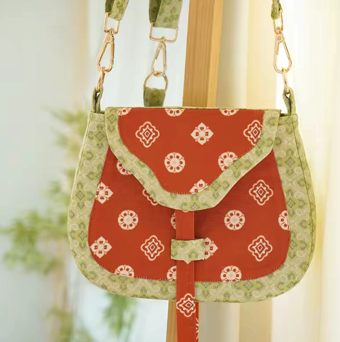 Full front view of a red and green Tang Dynasty floral Chinese canvas crossbody bag hanging on a wooden display stand, showing the saddle shape, all-over pattern, and gold-tone adjustable strap