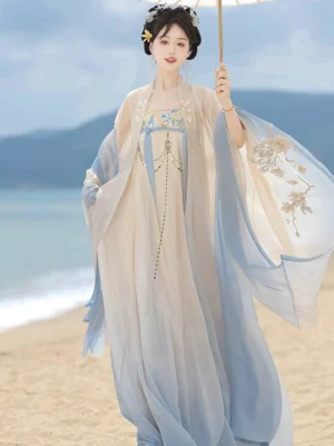 Tang Dynasty Hanfu Heziqun dress set with cream to sky blue gradient fabric, wide embroidered sleeves, chest tassel ornament, and flowing Pibo sash shown beside the seaside
