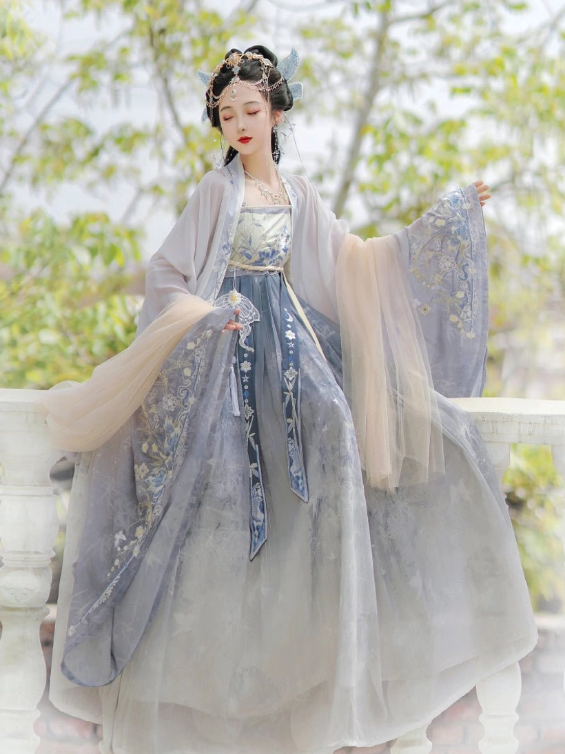 Blue Tang Dynasty Hanfu Heziqun dress set worn by model outdoors, featuring a beige wrap dress, flowing wide sleeve coat, embroidered waist sash and elegant layered silhouette