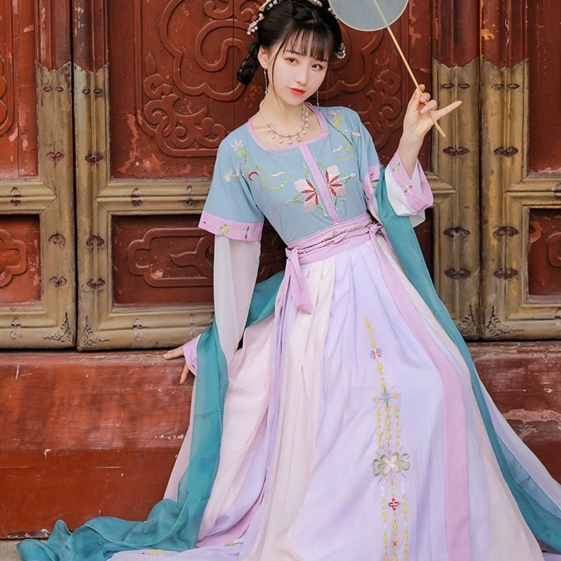 Three-quarter seated view of a lavender pink Tang-inspired Qiyao Ruqun Hanfu with vertical floral embroidery, small silk fan, and long cyan sash beside a carved red door
