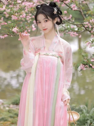 Front view of a Tang Dynasty Hanfu Qixiong Ruqun in a peach blossom garden with a pink gradient pleated skirt, pale pink embroidered top, green waist ribbons, and a wicker basket