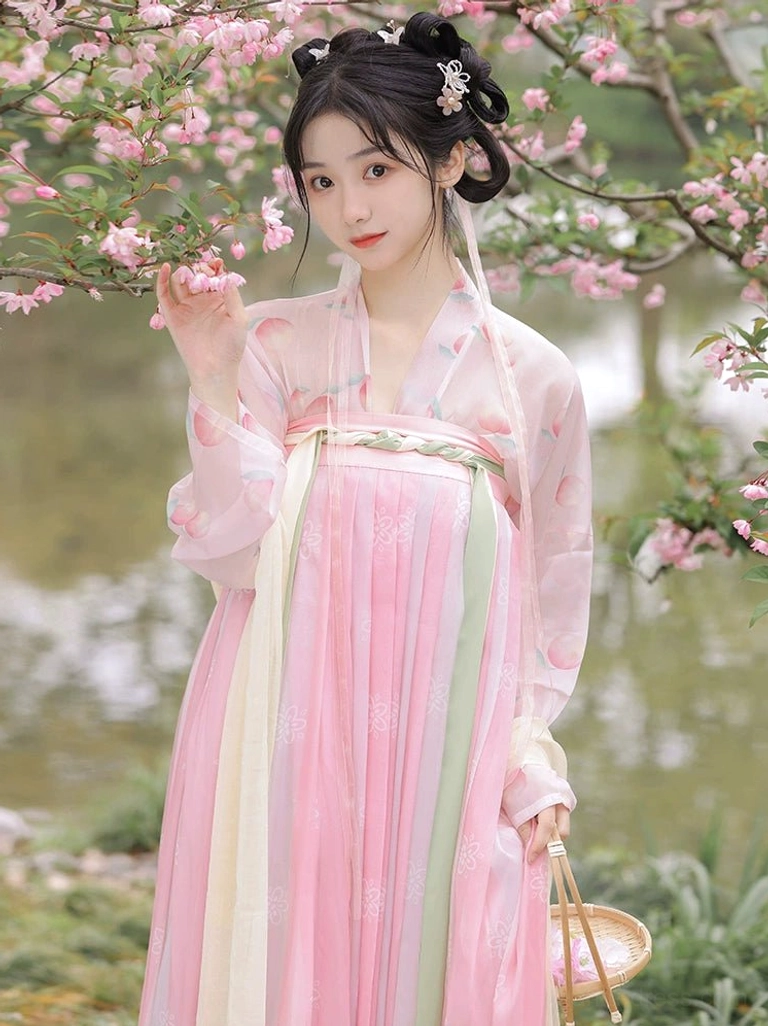 Front view of a Tang Dynasty Hanfu Qixiong Ruqun in a peach blossom garden with a pink gradient pleated skirt, pale pink embroidered top, green waist ribbons, and a wicker basket
