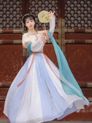 Three-quarter view of a cream yellow and pink Tang Dynasty Hanfu Qiyao Ruqun with floral embroidery, flowing cyan sash, and round silk palace fan beside a carved red wooden door